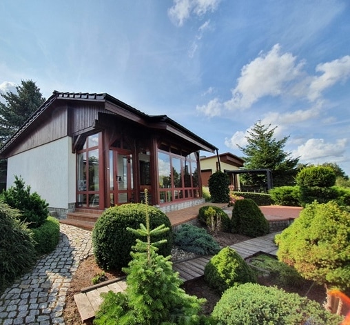 The exterior of the house features a large winter garden with expansive glass windows, surrounded by well-maintained greenery. A stone path leads through the landscaped garden, creating a serene approach to the entrance. The roofline presents a combination of modern and traditional architectural elements.