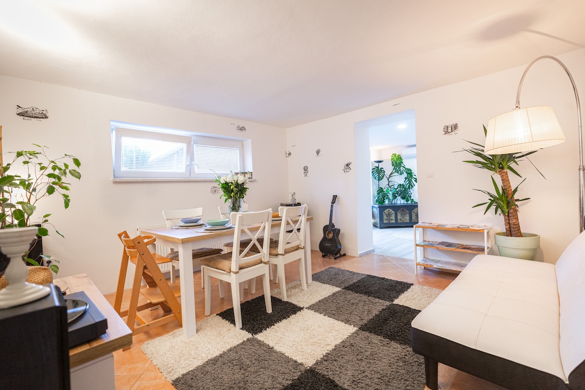 A cozy living area is illuminated by natural light from two windows, featuring a dining table surrounded by four chairs. An inviting rug defines the space, while greenery adds a touch of nature. A white sofa and floor lamp provide additional comfort and style.