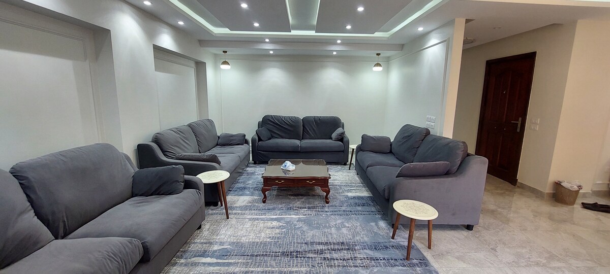 A spacious living area is furnished with multiple gray sofas arranged around a central wooden coffee table. A subtle area rug adds a soft touch, while recessed lighting enhances the bright and open atmosphere.