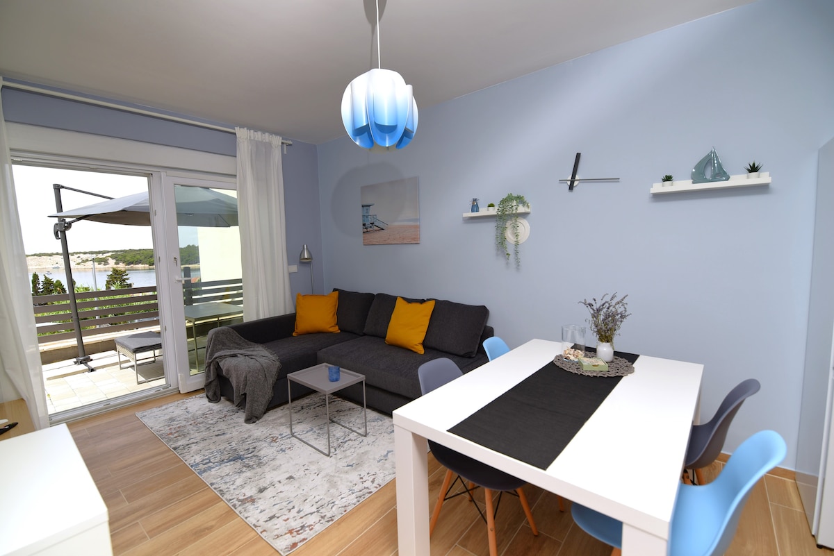 A modern living area is presented, featuring a dark sectional sofa adorned with yellow accent pillows. A dining table with colorful chairs accompanies a neutral rug on light wood flooring. Large glass doors open to a balcony, allowing natural light to fill the room.