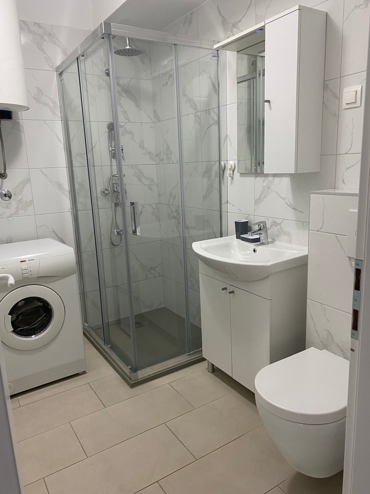 A modern bathroom is displayed, featuring a glass shower enclosure alongside a sleek white vanity with an integrated sink. A washing machine is positioned to the left, while a wall-mounted mirror enhances the space. Neutral tiles contribute to the clean and bright atmosphere.