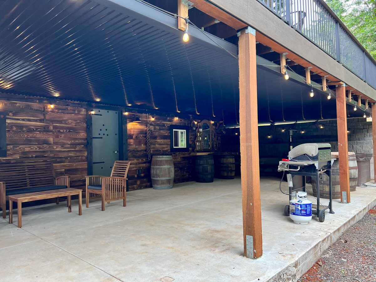 A spacious covered patio features wooden seating and decorative barrels, creating a relaxing outdoor environment. A barbecue grill is positioned nearby, and warm string lights hang from the ceiling, enhancing the ambiance for gatherings and meals.