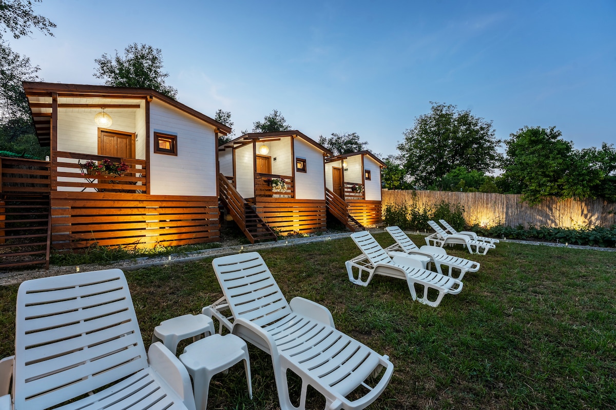 Three charming guest cabins are set against a serene evening backdrop, surrounded by a green lawn. Each cabin is well-lit, featuring wooden accents and steps leading up to the entrances. Several lounge chairs are placed invitingly on the grass, offering a peaceful space for relaxation.