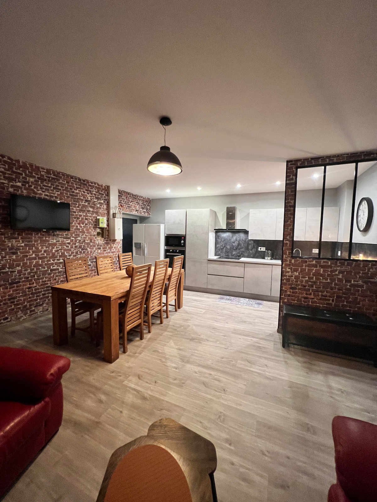 A modern dining area features a large wooden table surrounded by six wooden chairs, with a cozy red armchair nearby. The sleek kitchen showcases gray cabinetry and is separated by a glass partition, while exposed brick walls add texture to the space.