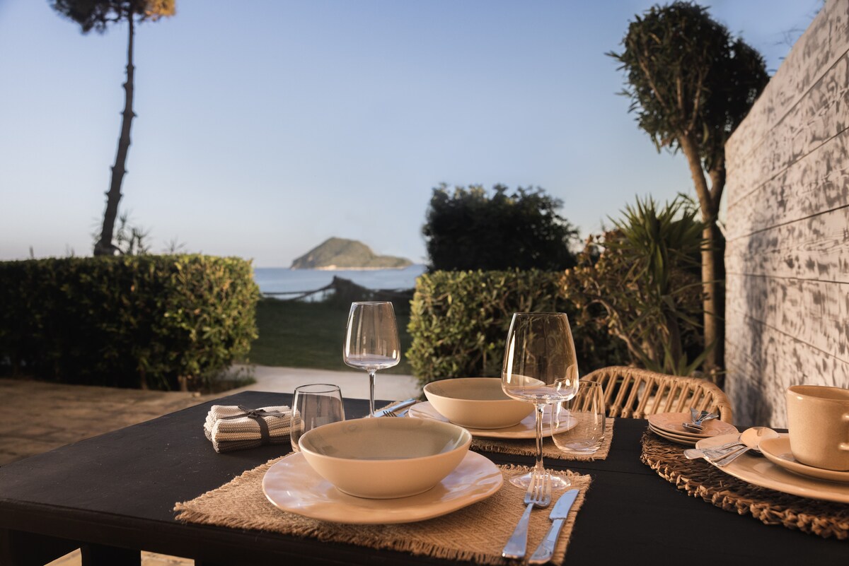 An outdoor dining area is presented with a table set for a meal, featuring plates, glasses, and utensils. Lush greenery surrounds the space, framed by a view of the sea and a distant island. Natural light enhances the tranquil atmosphere.