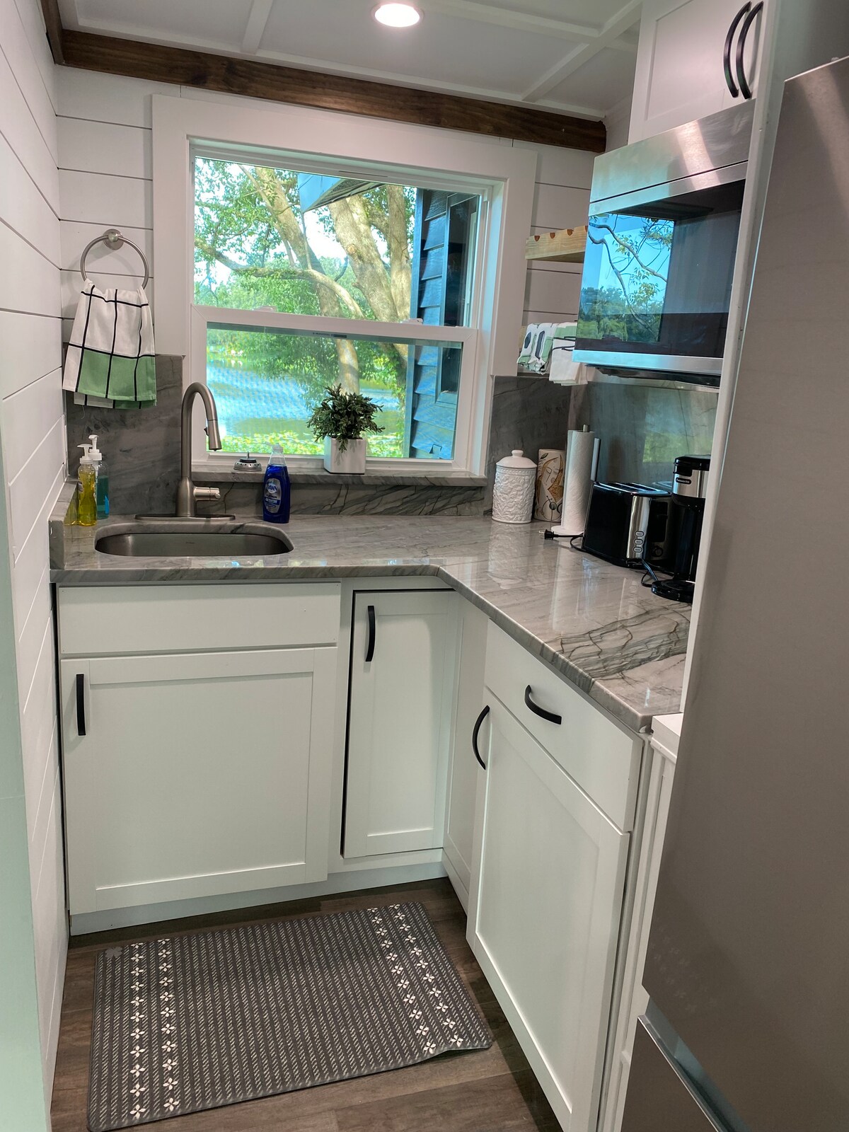 A small kitchen area is equipped with modern appliances and a sink, surrounded by white cabinetry and a granite countertop. A window offers a view of the outside greenery, adding natural light to the space. A textured mat lies underfoot.