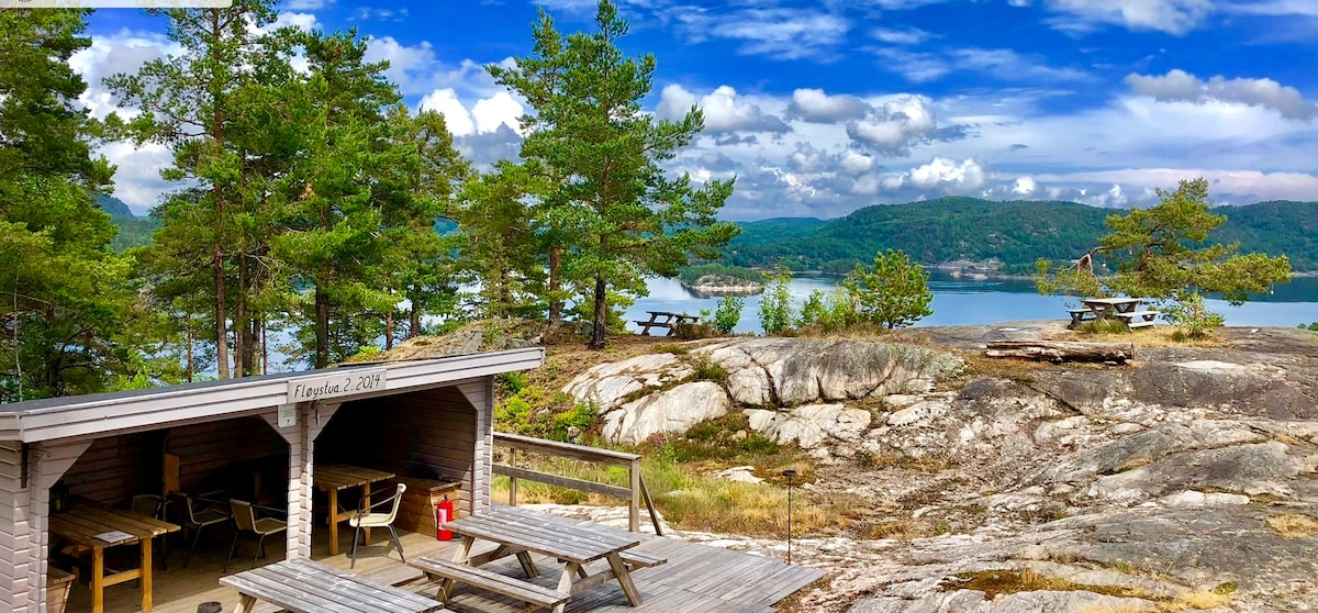 A wooden structure with picnic tables is set among green trees on rocky terrain. The view includes distant hills and calm water reflecting clouds, creating a serene outdoor space ideal for relaxation and social gatherings.