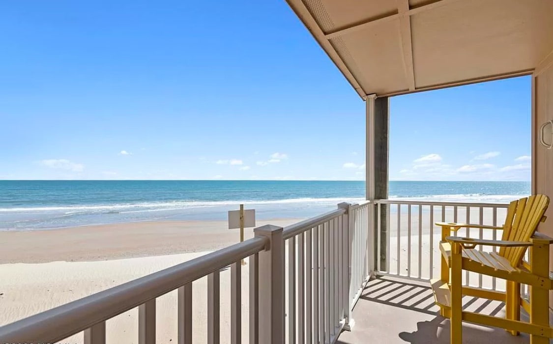 A private balcony overlooks the sandy beach and ocean, with a clear blue sky arching above. A bright yellow chair is positioned for relaxation, while the railing and flooring are painted in white, complementing the waterfront view.