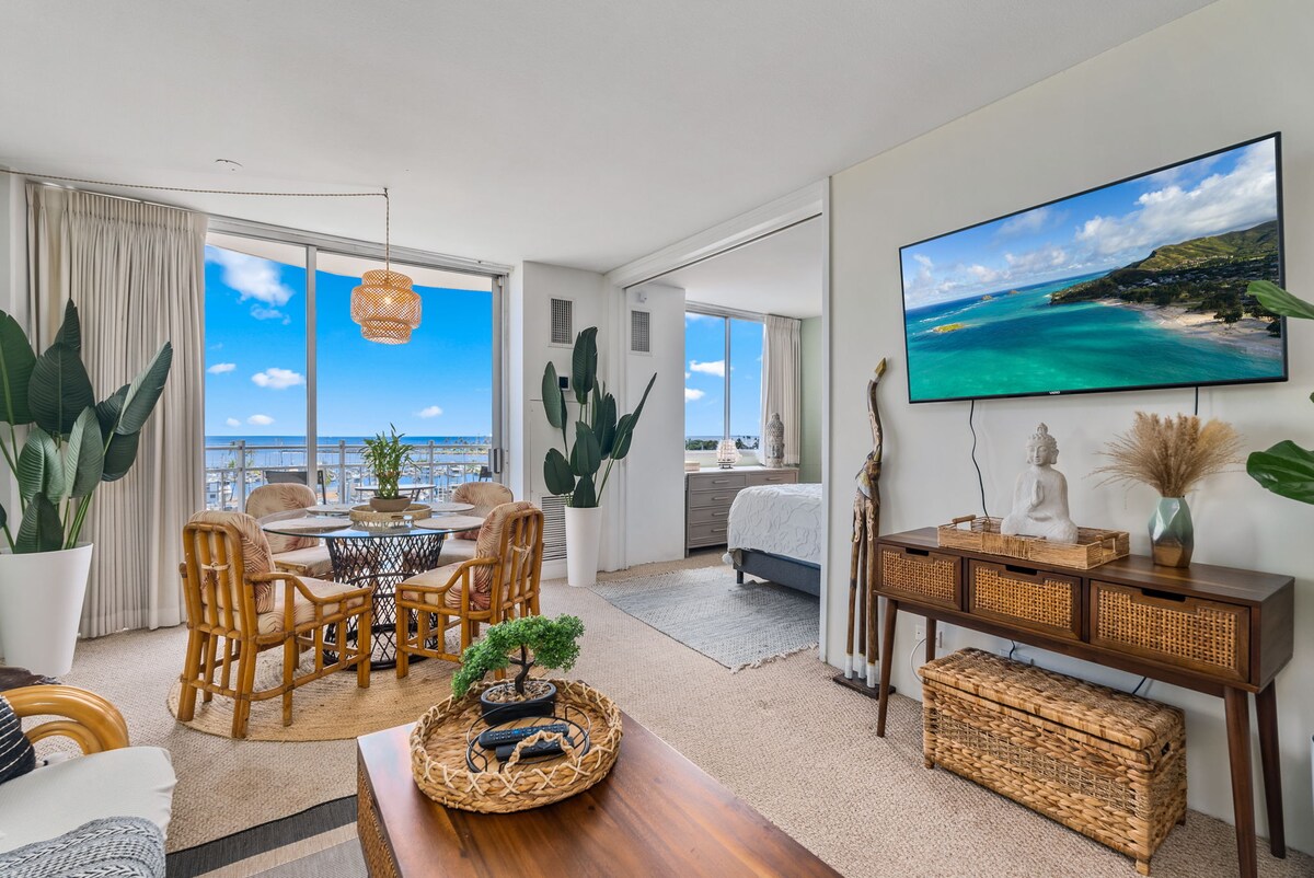 A bright living space features large windows with views of the ocean and beach. The dining area includes a round table surrounded by rattan chairs. A media console displays a large television, and decorative elements like plants and artwork enhance the inviting atmosphere.