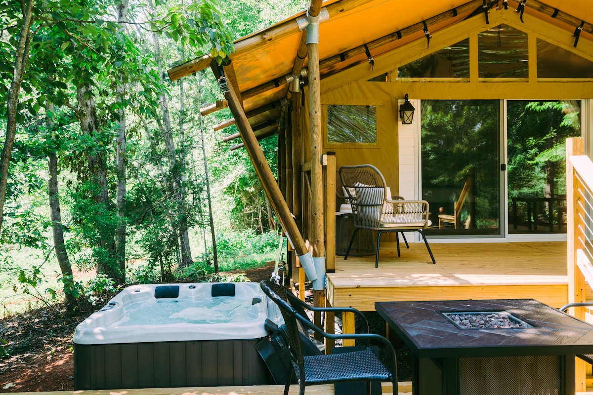 A hot tub is positioned on the deck next to a seating area, featuring a chair and a small table. The tent's entrance is visible through large glass doors, surrounded by lush greenery that provides a sense of privacy.