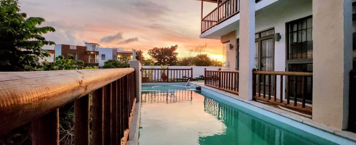 A tranquil scene is captured featuring a private pool reflecting the warm hues of a sunset. Balconies with wooden railings extend from the building, offering a scenic view of the lush garden and surrounding area.