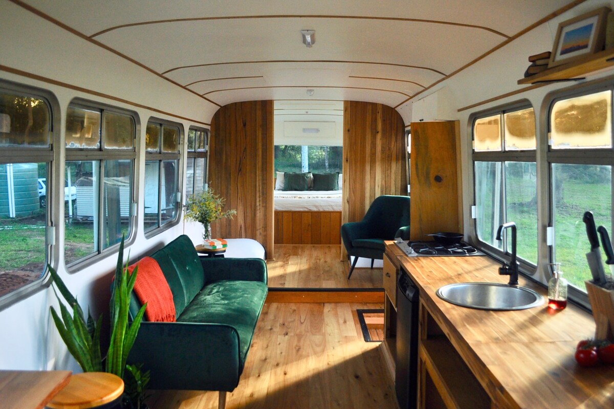 The interior of a restored bus features natural wood paneling, a spacious layout, and large windows allowing light to flood the space. A cozy sofa and armchair are positioned adjacent to a compact kitchen equipped with a sink and gas stovetop. A queen bed is visible in the rear area.