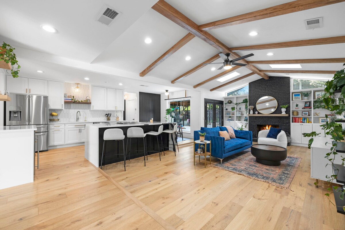 An open-concept living area features a modern kitchen with white cabinetry and stainless steel appliances. A blue sofa is centered around a circular coffee table, with a decorative rug beneath. Large windows invite natural light, and wooden beams add architectural interest.