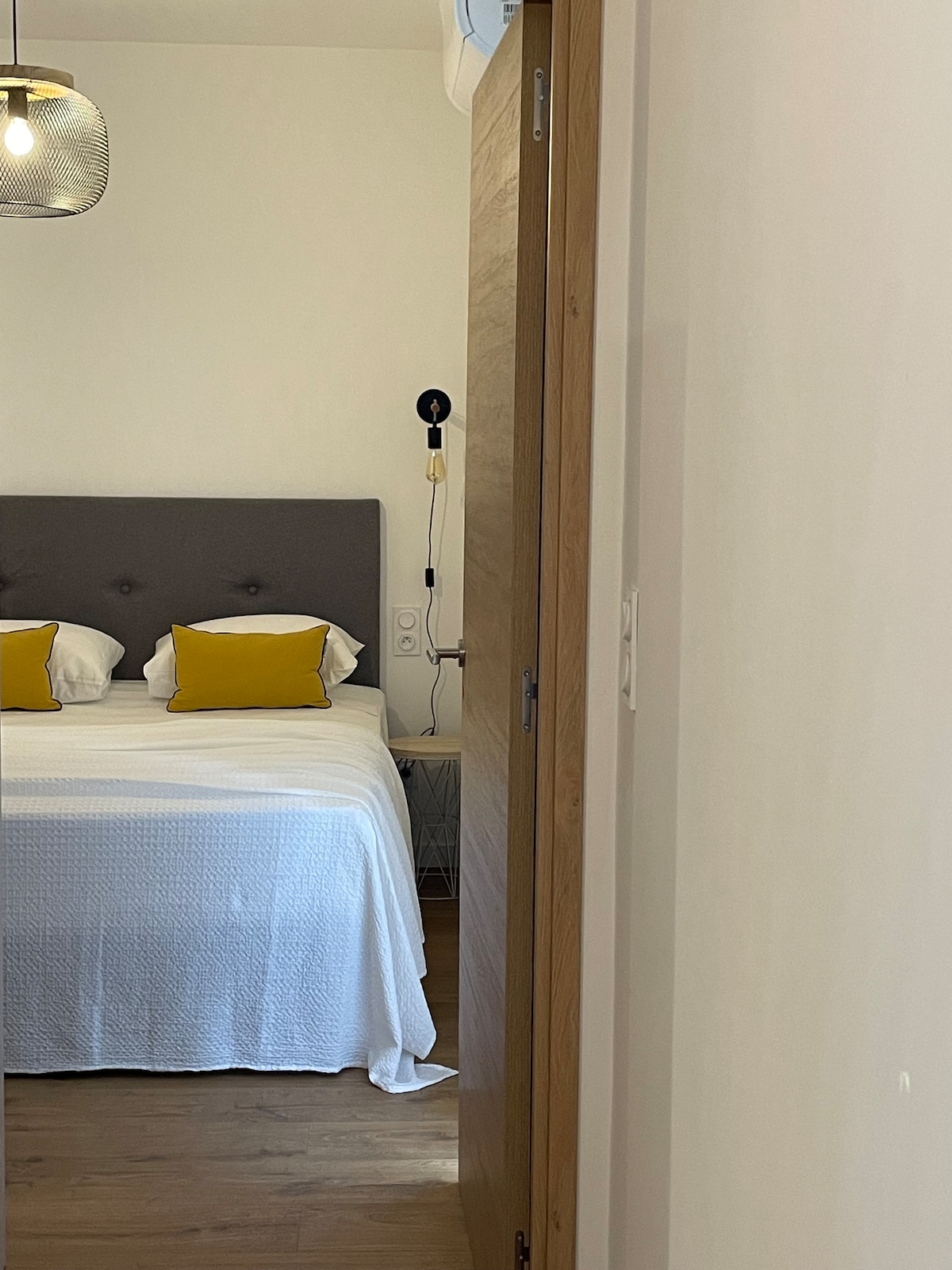 A view into a well-lit bedroom is presented, featuring a spacious bed adorned with white bedding and two yellow accent pillows. Wooden flooring and a subtle light fixture contribute to a cozy atmosphere, while a stylish door is slightly ajar, leading into the room.
