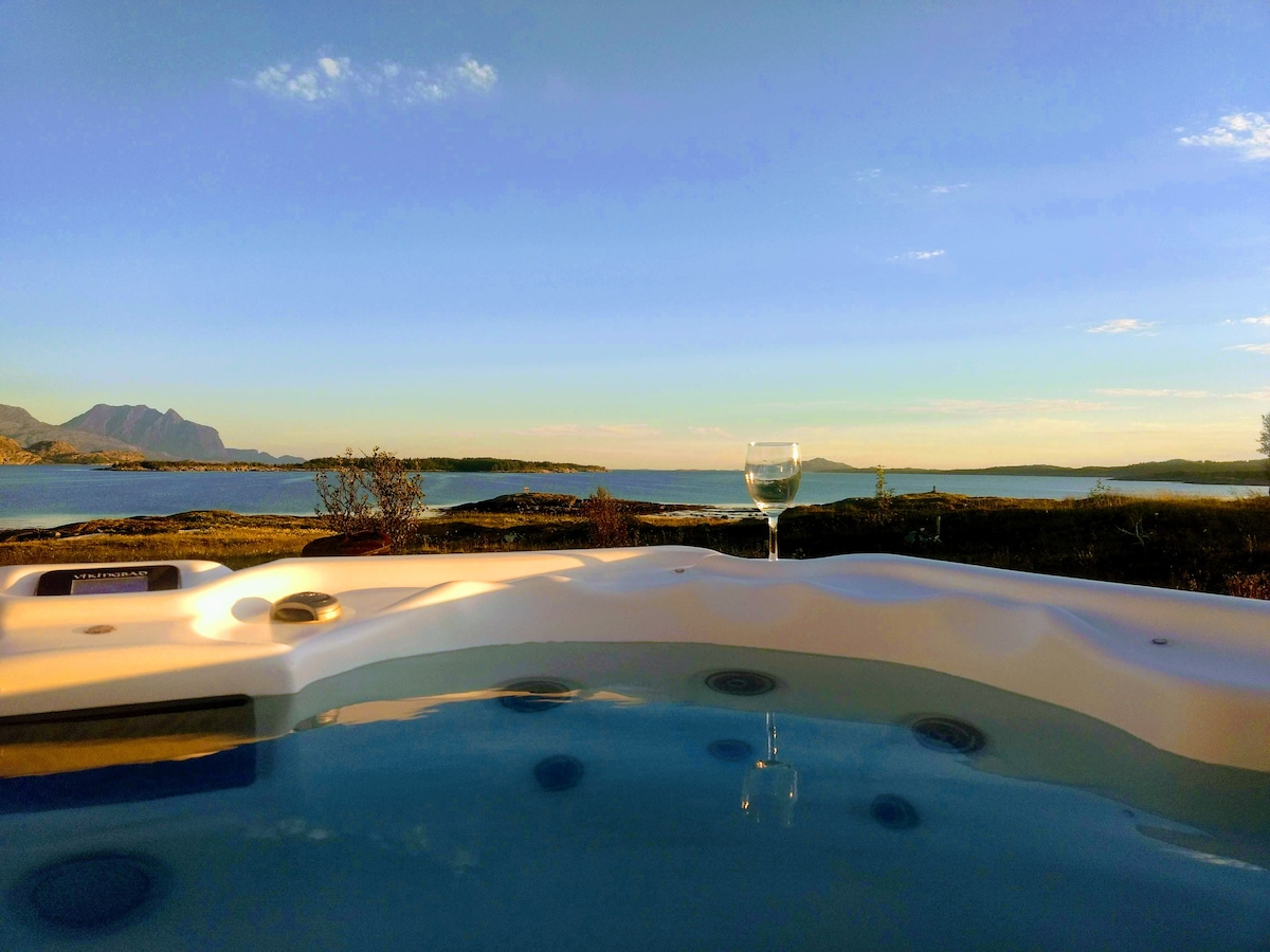 A jacuzzi is positioned on a terrace, overlooking a serene body of water. The view includes distant mountains under a clear sky, with soft evening light reflecting off the water's surface. A glass is placed on the edge of the jacuzzi, adding to the tranquil setting.