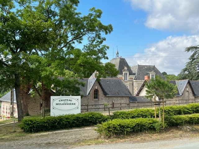 The entrance to Château Mulonnière is framed by lush greenery and features a well-maintained sign. The charming stone buildings of the gîte are visible, set against a backdrop of traditional architecture. The atmosphere exudes tranquility, inviting guests to explore the surrounding landscape.