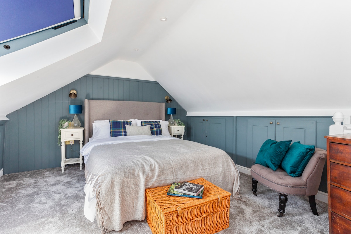 A bright and airy bedroom features a spacious bed with a light blanket and decorative pillows. Soft carpet covers the floor, complemented by a cozy armchair with teal cushions. Natural light fills the space through a skylight, and stylish bedside tables frame the bed.