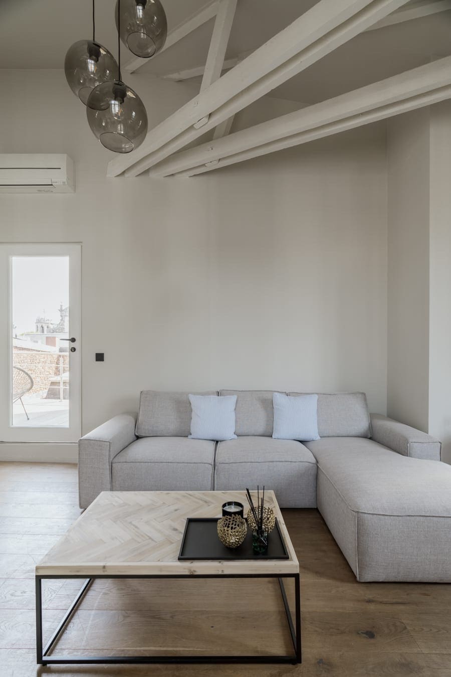 A light-filled living area features a gray sectional sofa adorned with soft pillows. A wooden coffee table with a tray sits in front. Large windows provide access to an outdoor terrace, while modern pendant lights hang from the white-beamed ceiling.