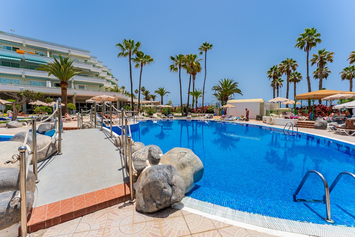 A spacious swimming pool is surrounded by tropical palm trees and rocks, inviting relaxation. Sun loungers are positioned along the poolside, and umbrellas provide shade. The bright blue water reflects the clear sky, emphasizing the serene outdoor environment.
