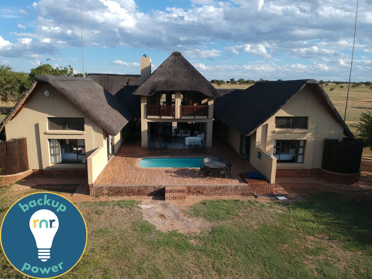 The holiday home features a thatched roof and a spacious patio area surrounding a private swimming pool. Large windows allow ample light into the interior. The scenic bush landscape is visible in the background, enhancing the outdoor setting.