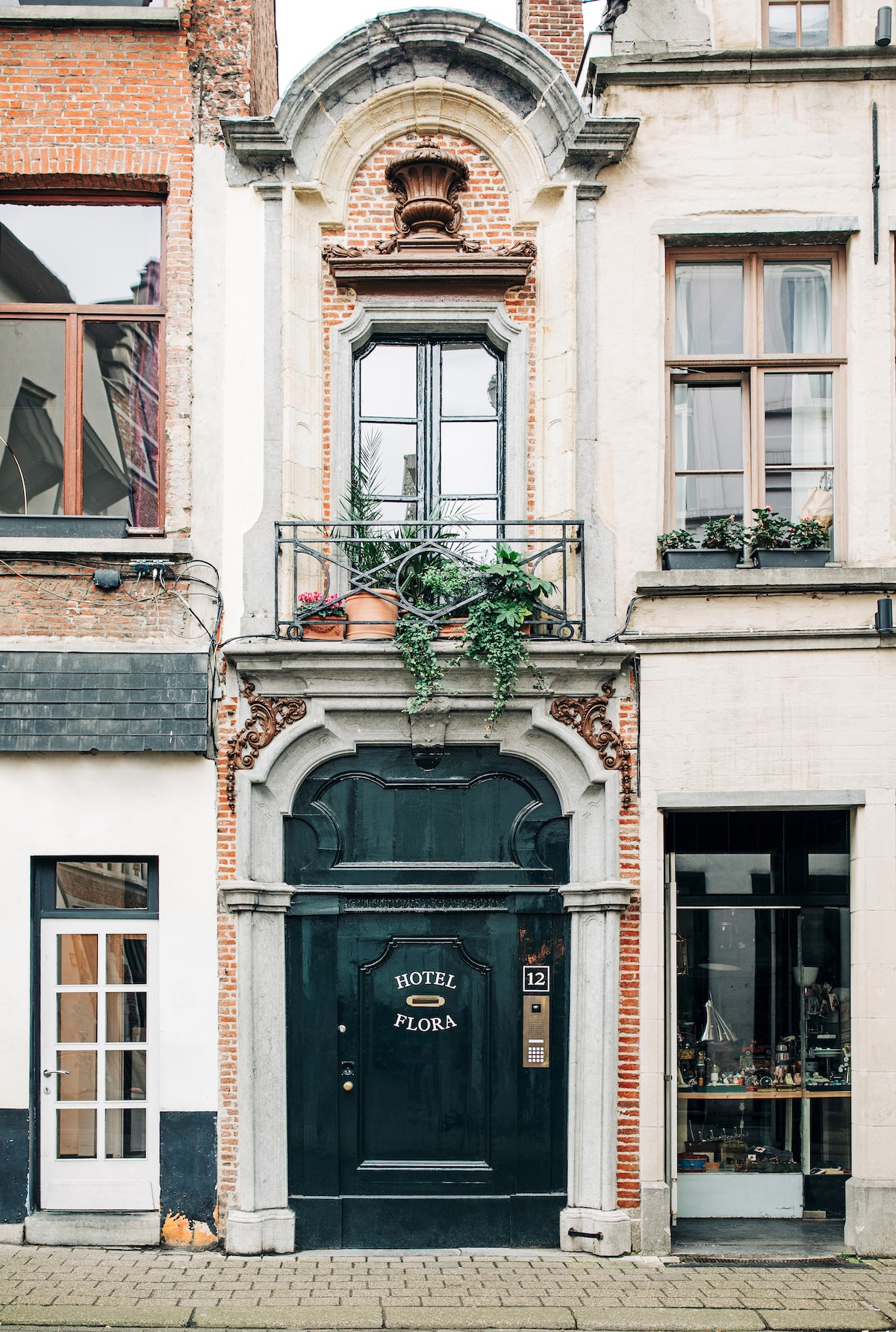 The entrance of Hotel Flora features a striking black door with the hotel name elegantly displayed. The ornate façade showcases architectural details, including a decorative arch and a small balcony adorned with greenery. Large windows on either side offer glimpses of the vibrant street.