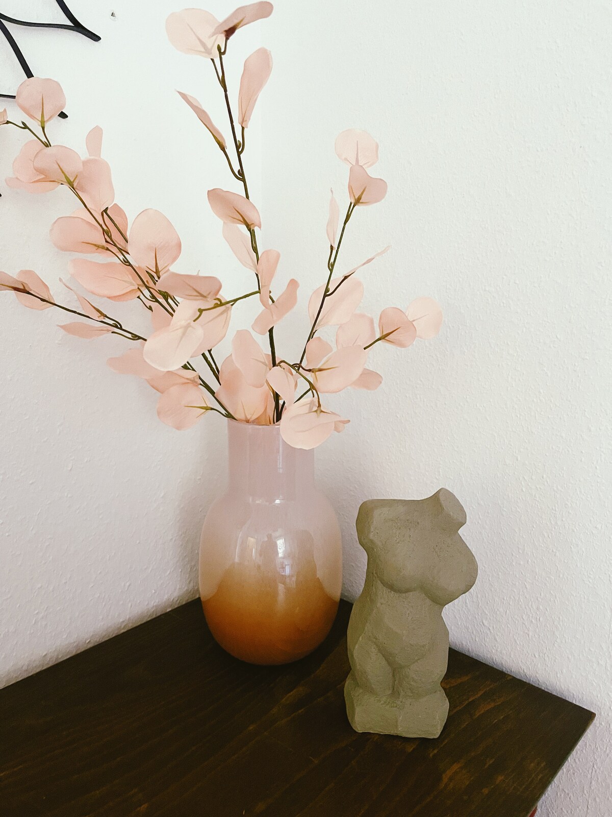 A decorative corner features a soft pink vase containing delicate foliage, complemented by a sculptural figure made of a natural material. The items rest on a dark wooden surface against a light wall, creating a serene focal point.