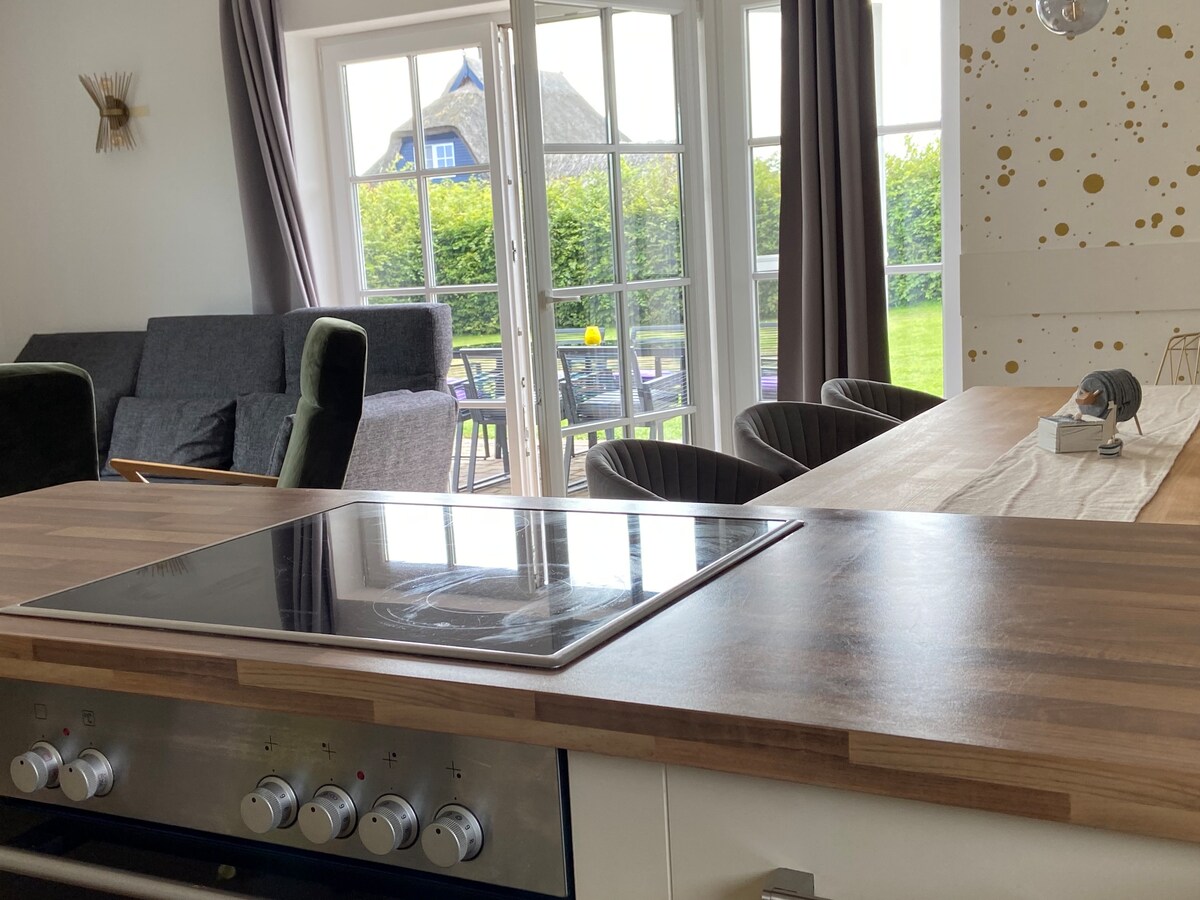 A modern kitchen with a wooden countertop is featured, highlighting a minimalist stove. An inviting living area is visible in the background, along with large glass doors leading to a garden space. Comfortable seating and a dining table are also included in the scene.