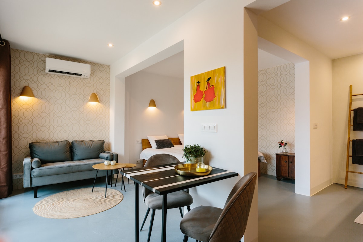 A modern studio is presented with a cozy seating area, featuring a gray sofa and small round table. Bright lighting enhances the space, while artwork adorns the walls. An inviting sleeping area is visible in the background, characterized by a comfortable bed, contributing to a serene ambiance.