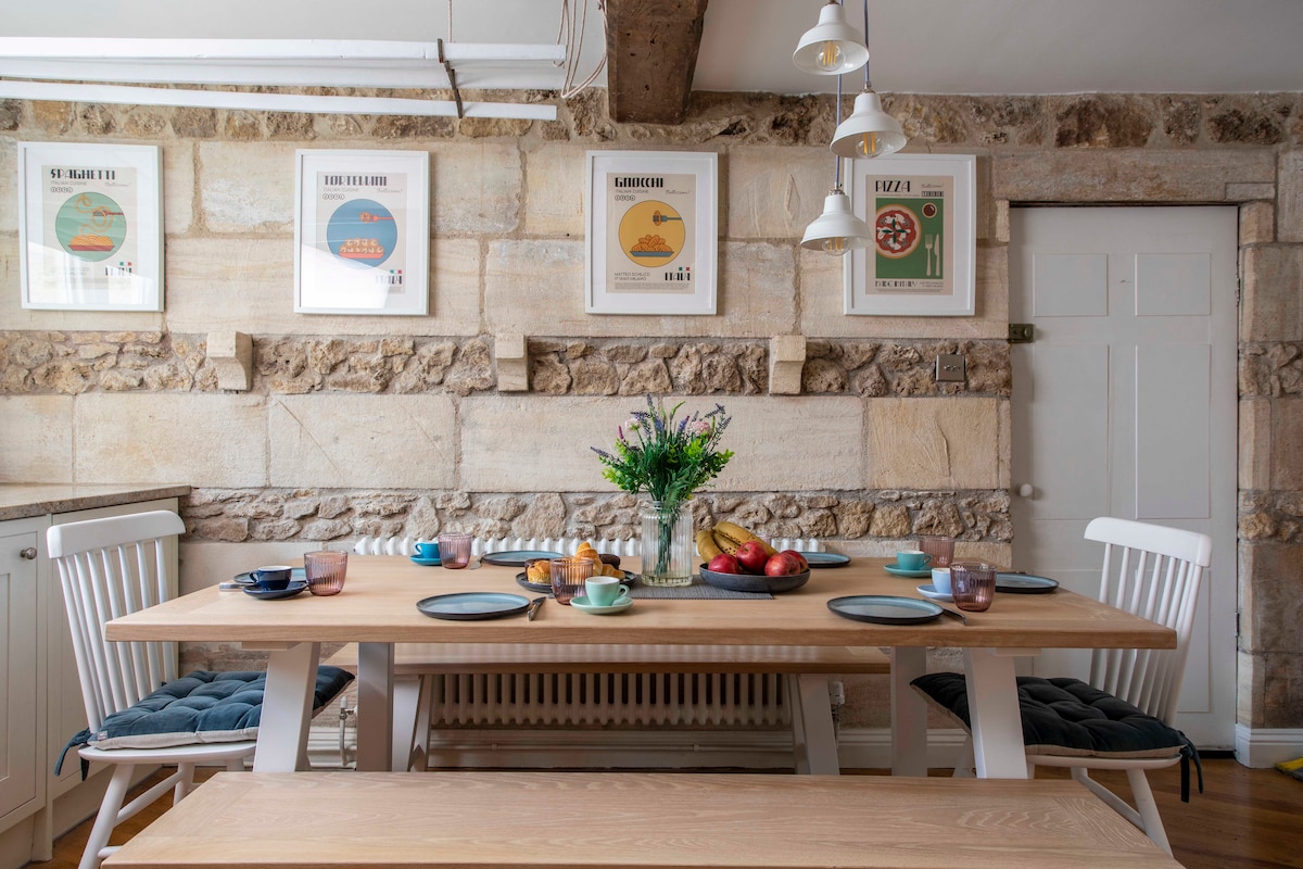A dining area features a long wooden table set with colorful dishes and fresh fruit. Natural stone walls provide texture, while framed artwork adds interest. Soft overhead lighting and a small vase of flowers enhance the inviting atmosphere.