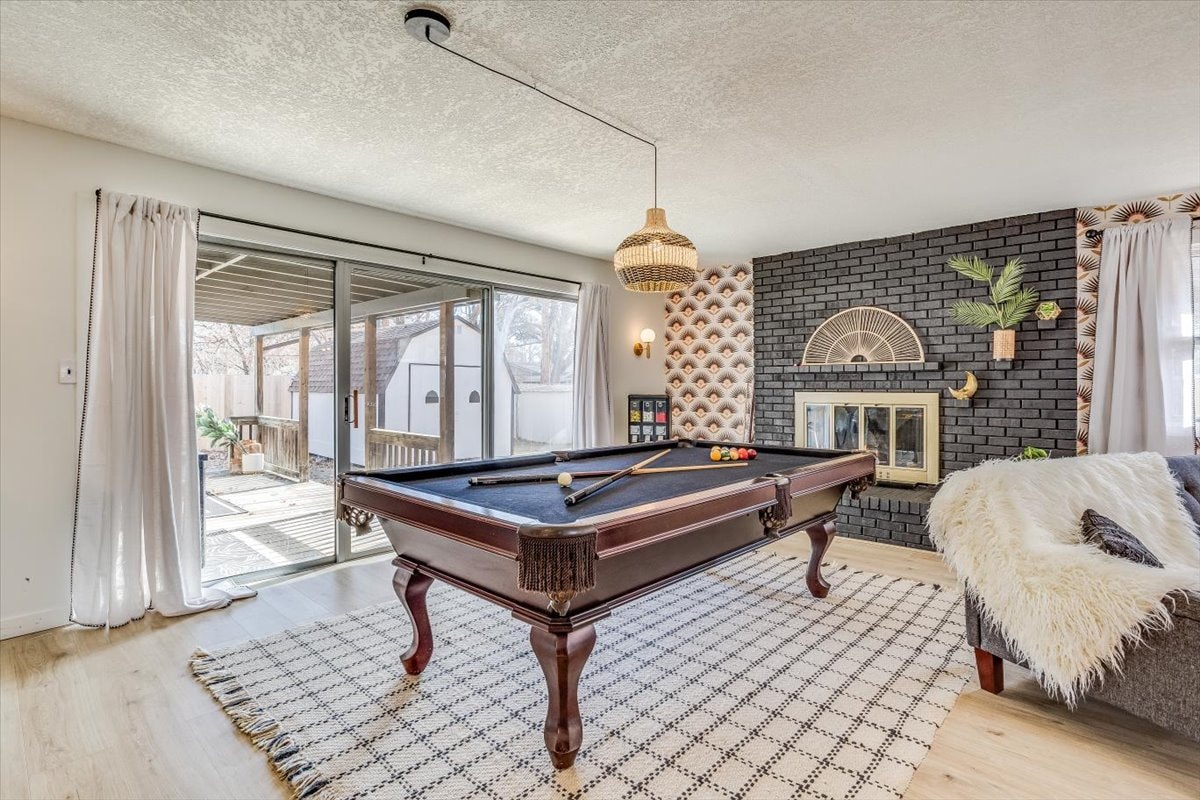 A spacious living area features a classic billiard table in the center, surrounded by natural light from large sliding doors. A cozy seating arrangement is nearby, complemented by a textured rug, while a decorative fireplace adds character to the room.
