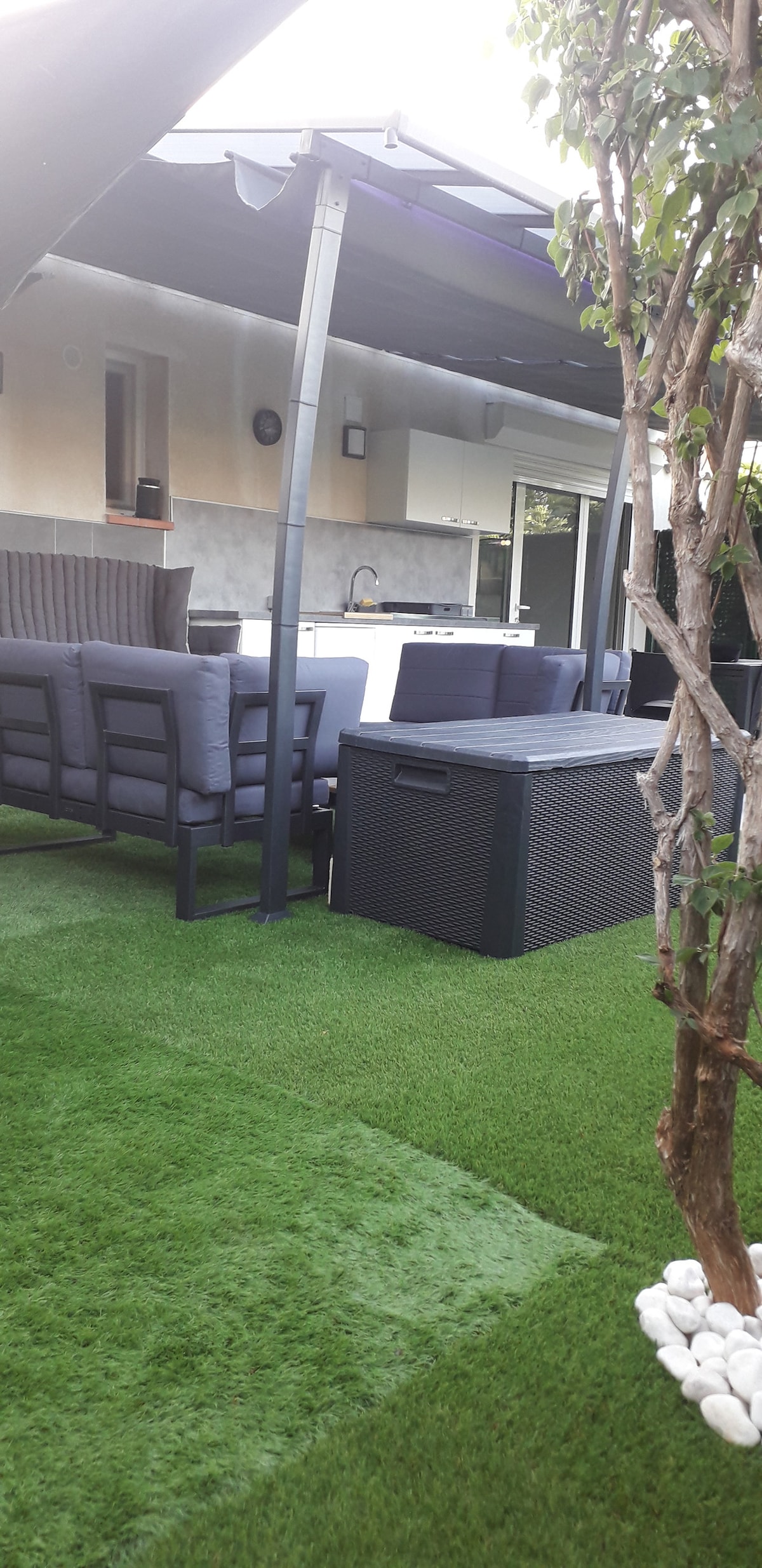 An outdoor area features a shaded seating arrangement with grey cushioned chairs surrounding a dark rectangular table. A small storage unit is visible, and lush green artificial grass completes the serene setting. A modern kitchen is partially seen in the background.