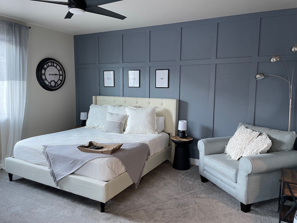 A light and airy sleeping area features a spacious bed with decorative pillows against a modern blue wall. A cozy armchair is positioned nearby, accompanied by a stylish lamp and a small nightstand. Natural light filters through sheer curtains, creating a calm ambiance.