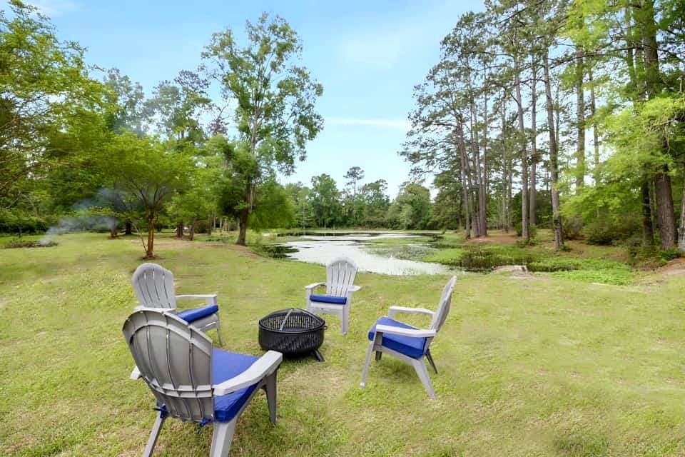 A spacious outdoor area is showcased with several chairs arranged around a fire pit, overlooking a serene pond. Lush greenery and tall trees create a peaceful backdrop, while the gently sloping lawn leads down to the water's edge.