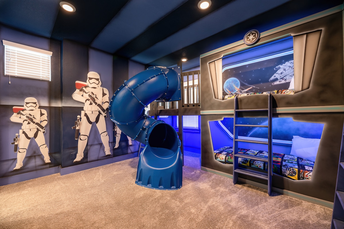 A themed children's bedroom featuring a blue slide leading from a bunk bed. Colorful bedding depicting planets is visible underneath the bed, while wall art includes figures resembling space soldiers. Soft carpeting and blue lighting contribute to a playful atmosphere.