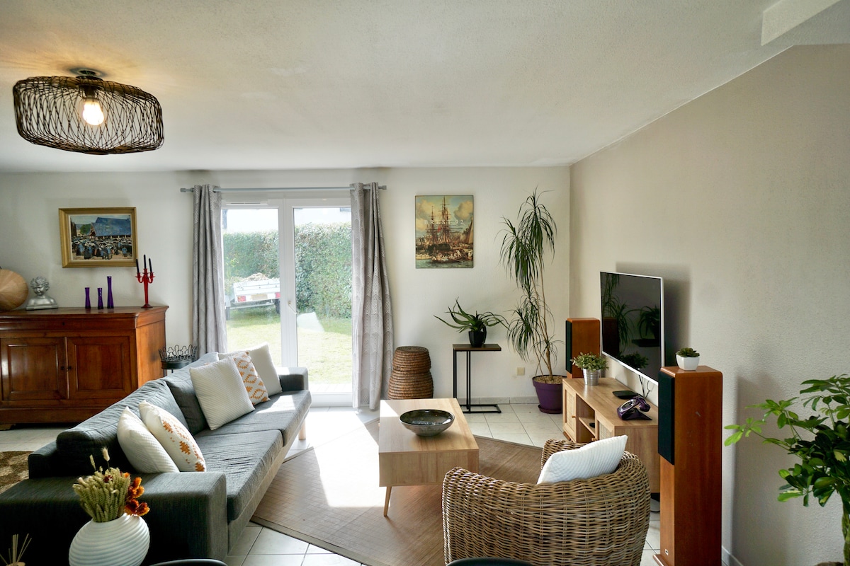 The living area includes a comfortable grey sectional sofa and light-colored armchairs surrounding a wooden coffee table. A flat-screen television is positioned on a unit against the wall. Large glass doors lead to the garden, allowing natural light to fill the space.