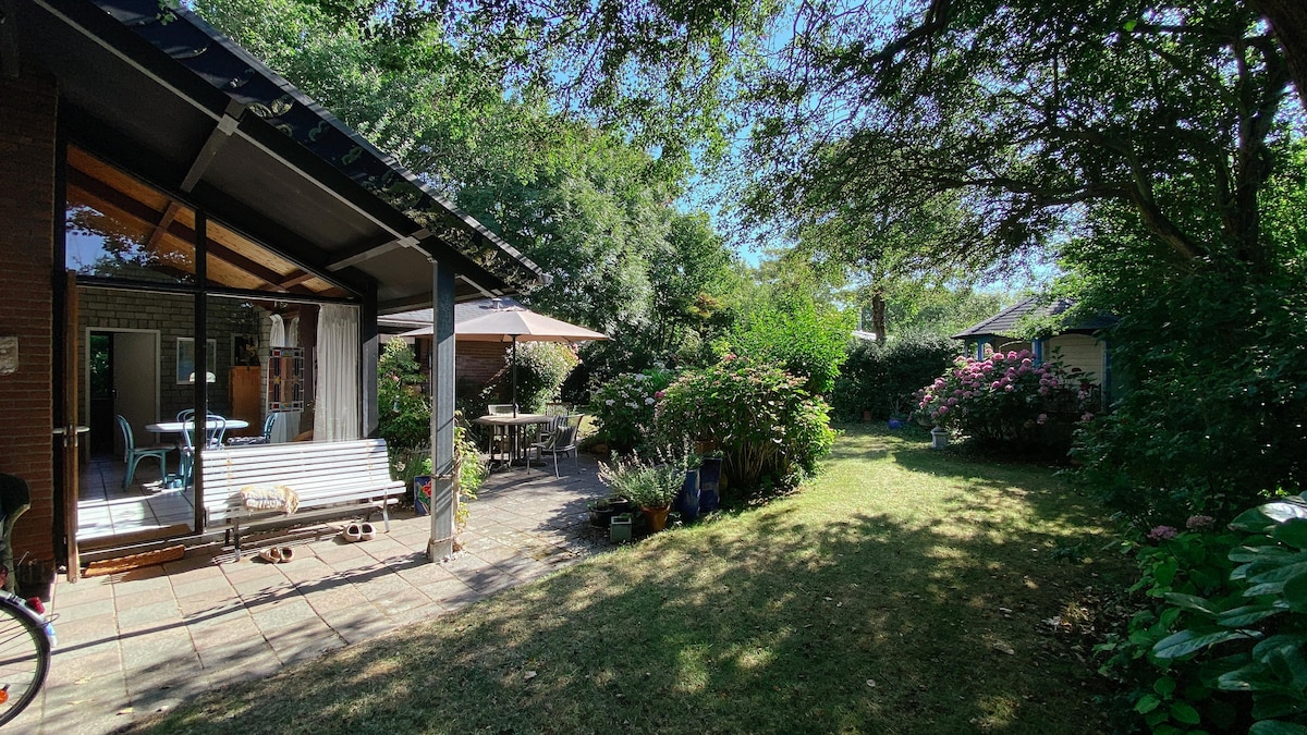 A serene garden is surrounded by vibrant greenery, featuring shaded seating areas and a patio table under an umbrella. The cozy house is visible through large glass doors, allowing light to fill the inviting interior space.