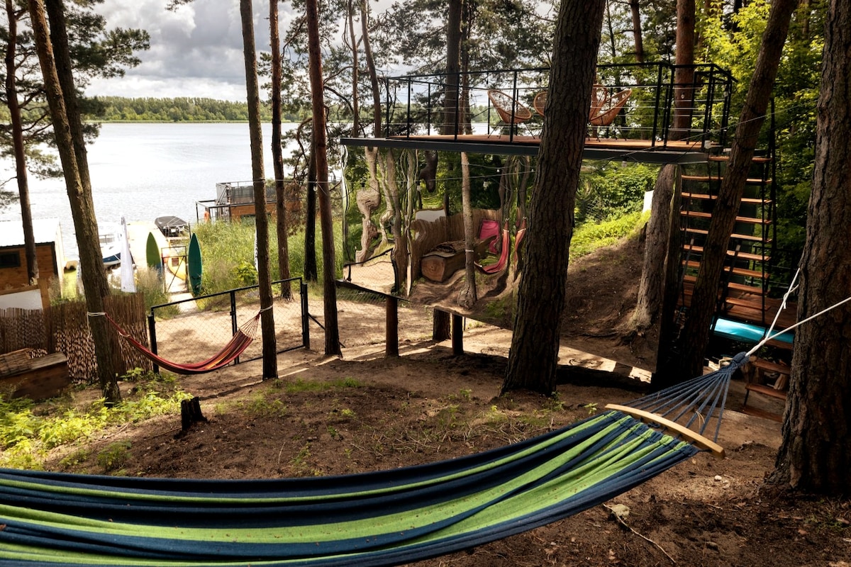 A serene outdoor space features a multicolored hammock nestled between trees, surrounded by lush greenery. A wooden deck above includes seating and is connected by stairs, leading to a calm lakeside area with boats visible in the background.