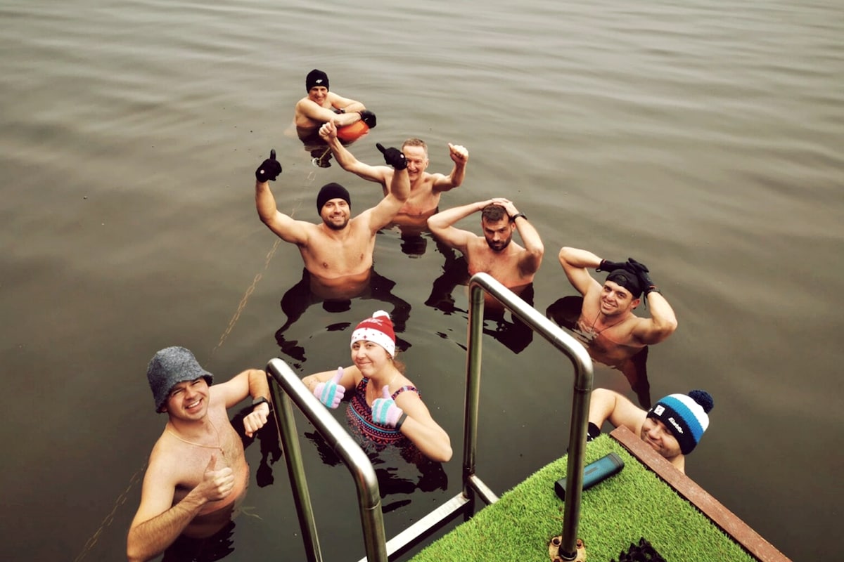 A group of individuals is enjoying a swim in a calm body of water. The scene captures a small gathering, with some participants standing in water while others are seated. A ladder leading from a dock is visible in the foreground.