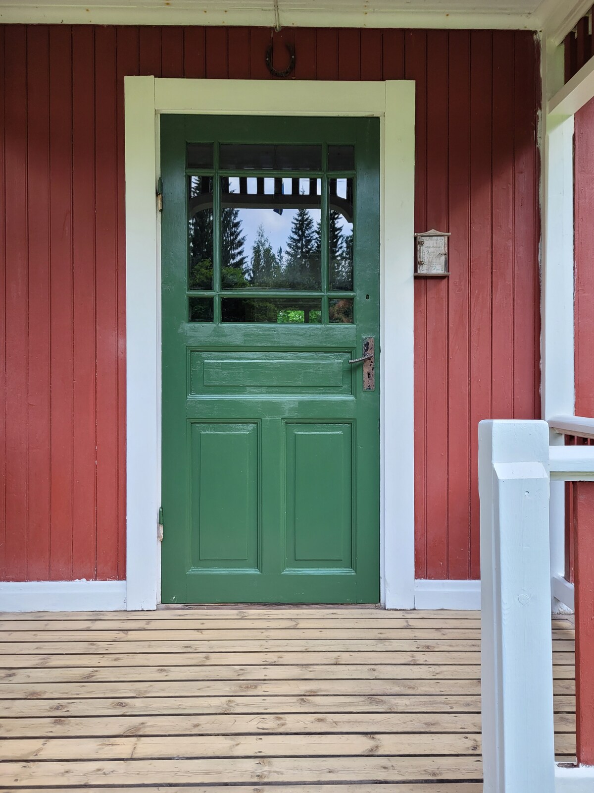 A green wooden door is framed by white trim, set against a vibrant red wooden exterior. The door features a small window with a grid pattern and is complemented by a wooden porch with a light-colored railing, enhancing the inviting entrance.