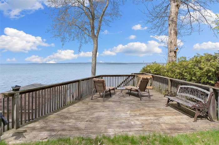 A spacious wooden deck overlooks the water, providing a serene outdoor space. Two lounge chairs are positioned for relaxation, while a decorative bench adds a cozy touch. Lush greenery is visible at the edge, enhancing the natural surroundings.