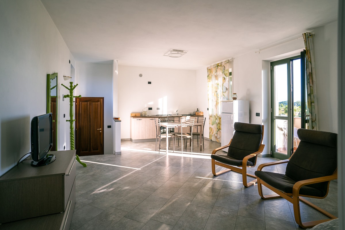 A light-filled living space is captured, featuring two wooden lounge chairs positioned beside a low dresser. A dining table with chairs is visible in the background, complemented by a kitchen area with modern appliances. Large windows offer views of the surrounding landscape.