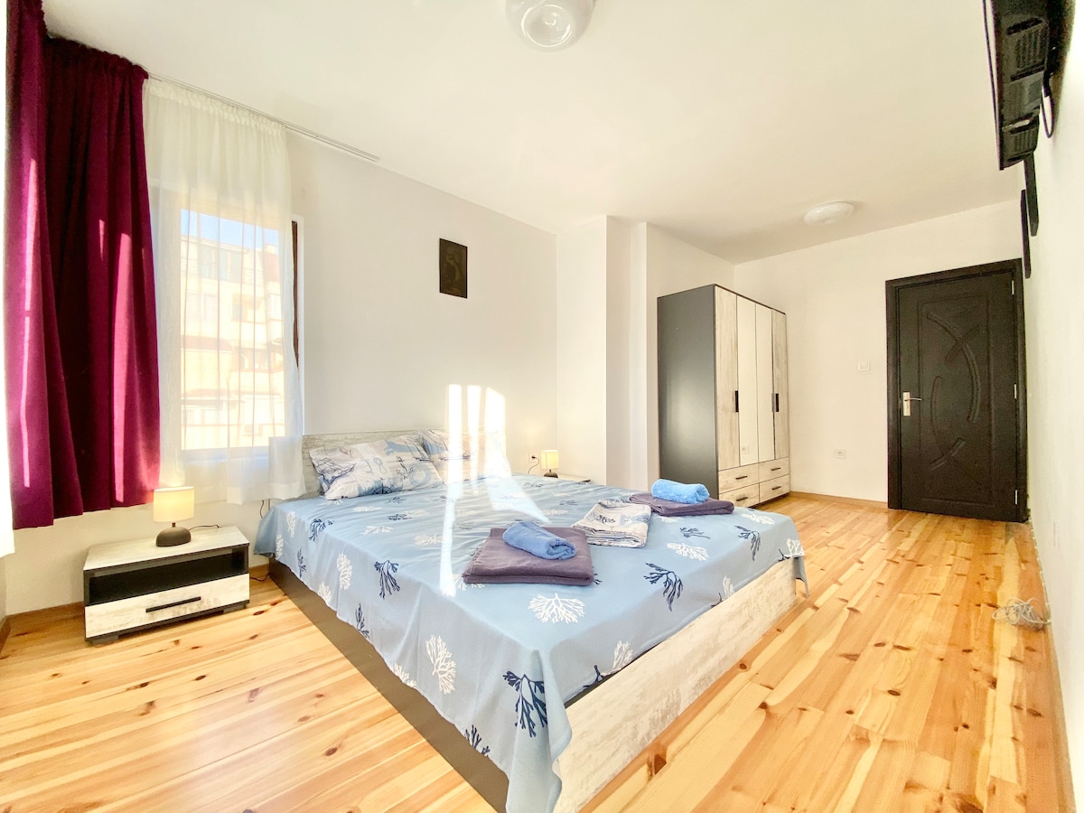 A spacious bedroom is depicted, featuring a large bed layered with soft linens. Natural light filters through sheer curtains, illuminating the wooden floor. A comfortable nightstand and a wardrobe complete the space, with towels neatly arranged at the foot of the bed.