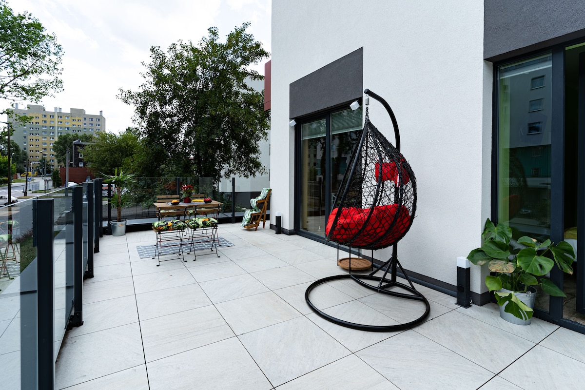 A spacious terrace is presented with large, light-colored tiles, complemented by a hanging chair featuring red cushions. A wooden table surrounded by chairs offers dining possibilities, while potted plants add greenery. Tall glass railings enhance safety and provide unobstructed views of the surroundings.