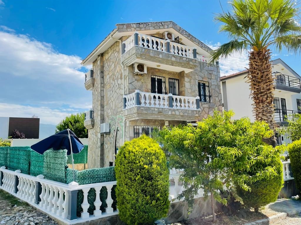 A multi-level villa constructed with stone accents is surrounded by lush greenery and palm trees. Balconies adorned with railings provide outdoor space for relaxation. A shaded area features an umbrella, offering a cool retreat next to the garden. The vibrant blue sky enhances the serene atmosphere.