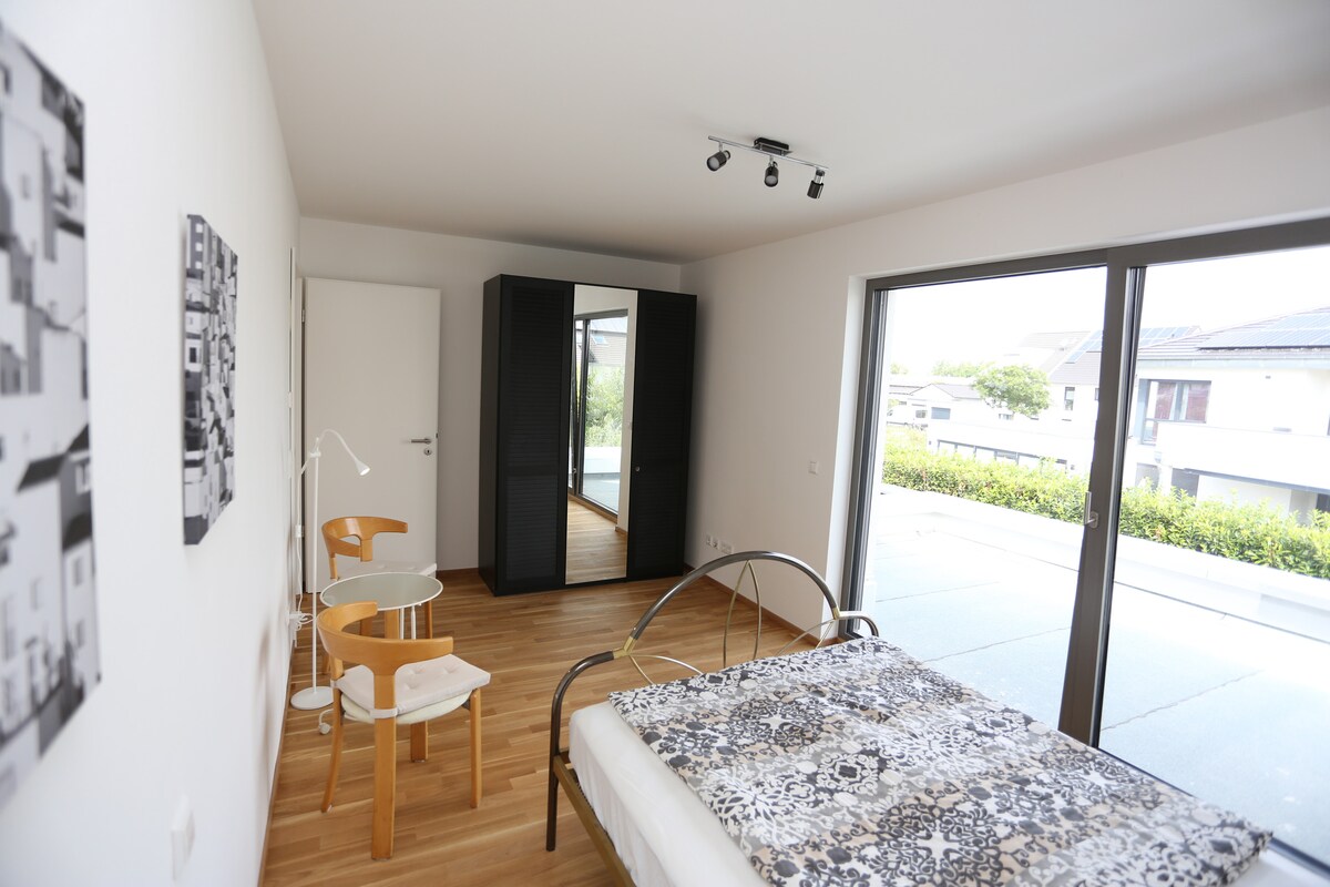 A bright bedroom features large glass sliding doors leading to a patio. The space includes a spacious bed with a patterned blanket, a modern wardrobe, and two wooden chairs. Natural light fills the room, highlighting the warm wooden flooring.