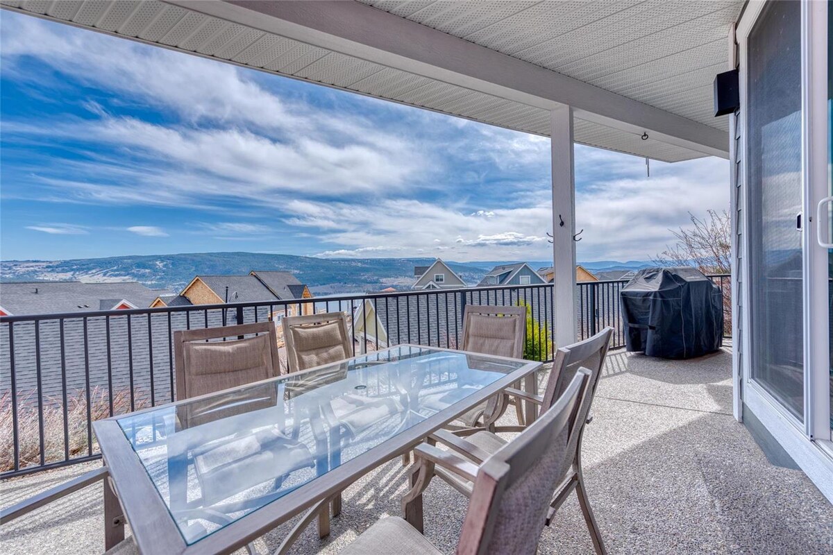 An outdoor patio features a glass-top dining table surrounded by six chairs, set against a backdrop of expansive lake and hillside views. A barbecue grill is positioned nearby, and the space is covered by an overhang for shade.