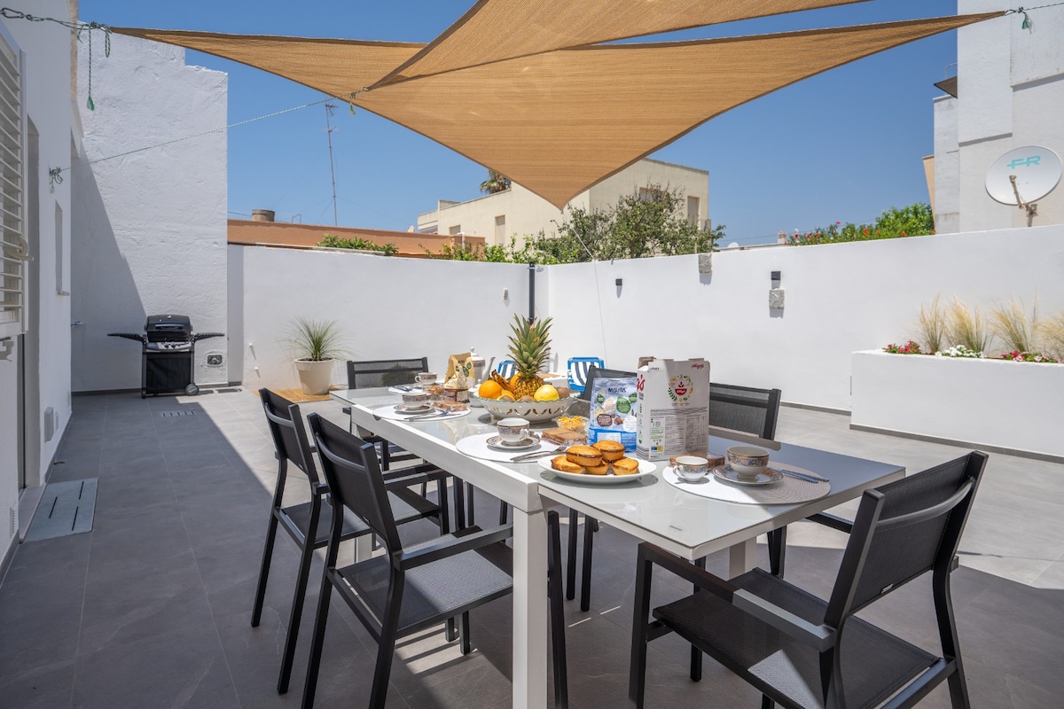 A spacious outdoor courtyard features a large dining table set for breakfast, including plates of fruit and pastries. A sunshade provides coverage from direct sunlight, and a barbecue grill is visible in the background. The area is surrounded by clean white walls and decorative plants.