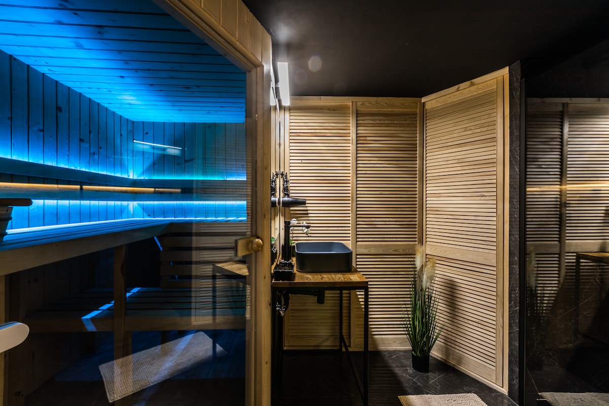 A modern sauna room features a wooden-paneled seating area illuminated by soft blue lighting. A sleek black sink is positioned beside a decorative plant, while sleek glass and wooden screens create a sense of privacy within the space.
