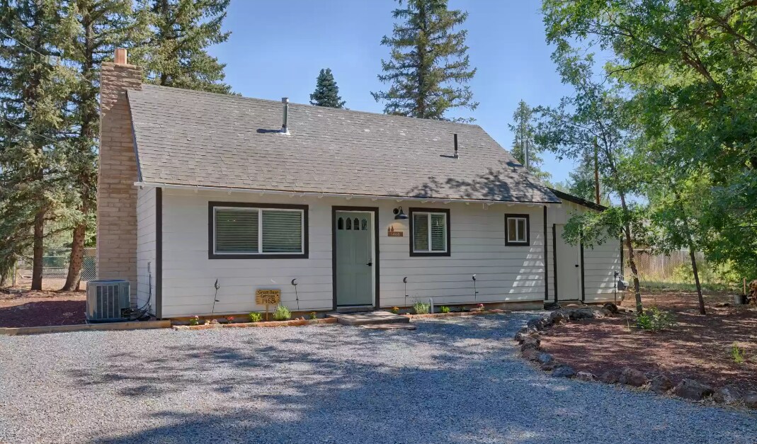 A charming exterior of a single-story home is observed, framed by tall trees. The house features a gravel driveway leading to a welcoming entrance, partially shaded by natural foliage. Windows are positioned symmetrically, allowing natural light to enter the interior.