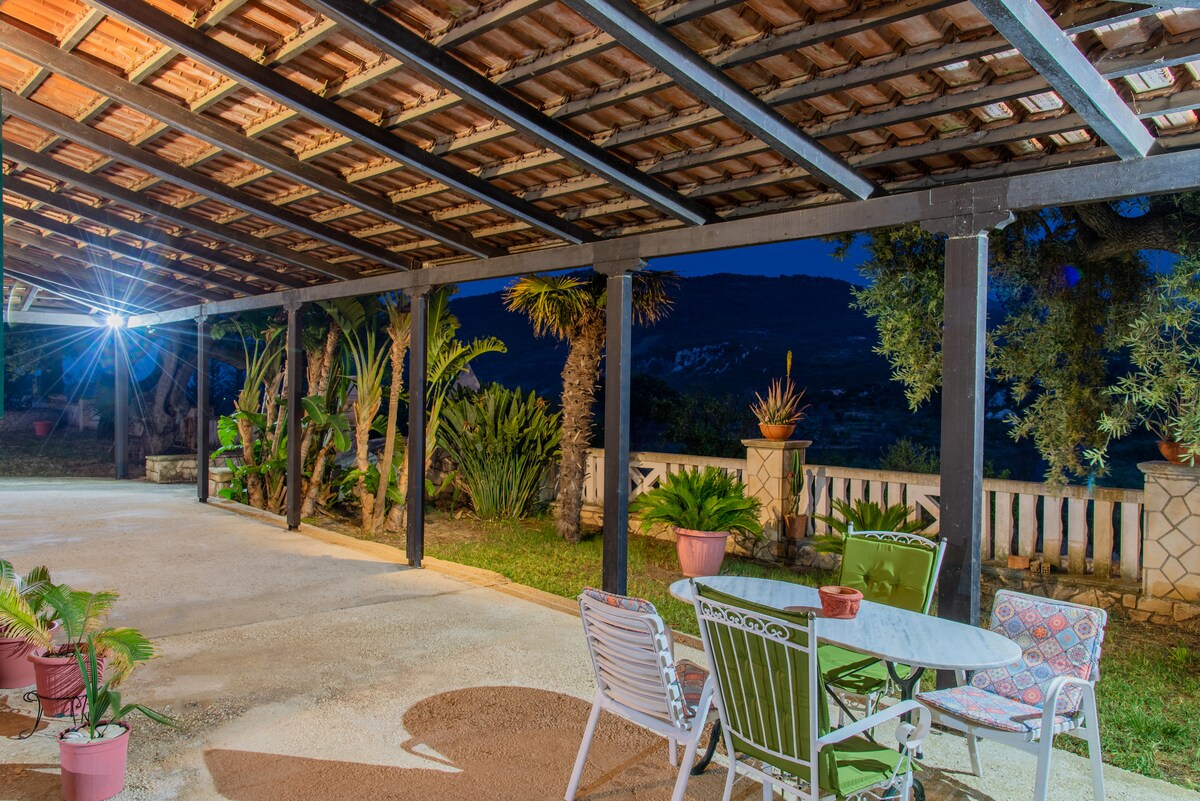 The outdoor area features a covered patio with a dining table and chairs, surrounded by lush greenery. Potted plants add touches of color, and the open space is well-lit, providing a comfortable setting for relaxation in the evenings.